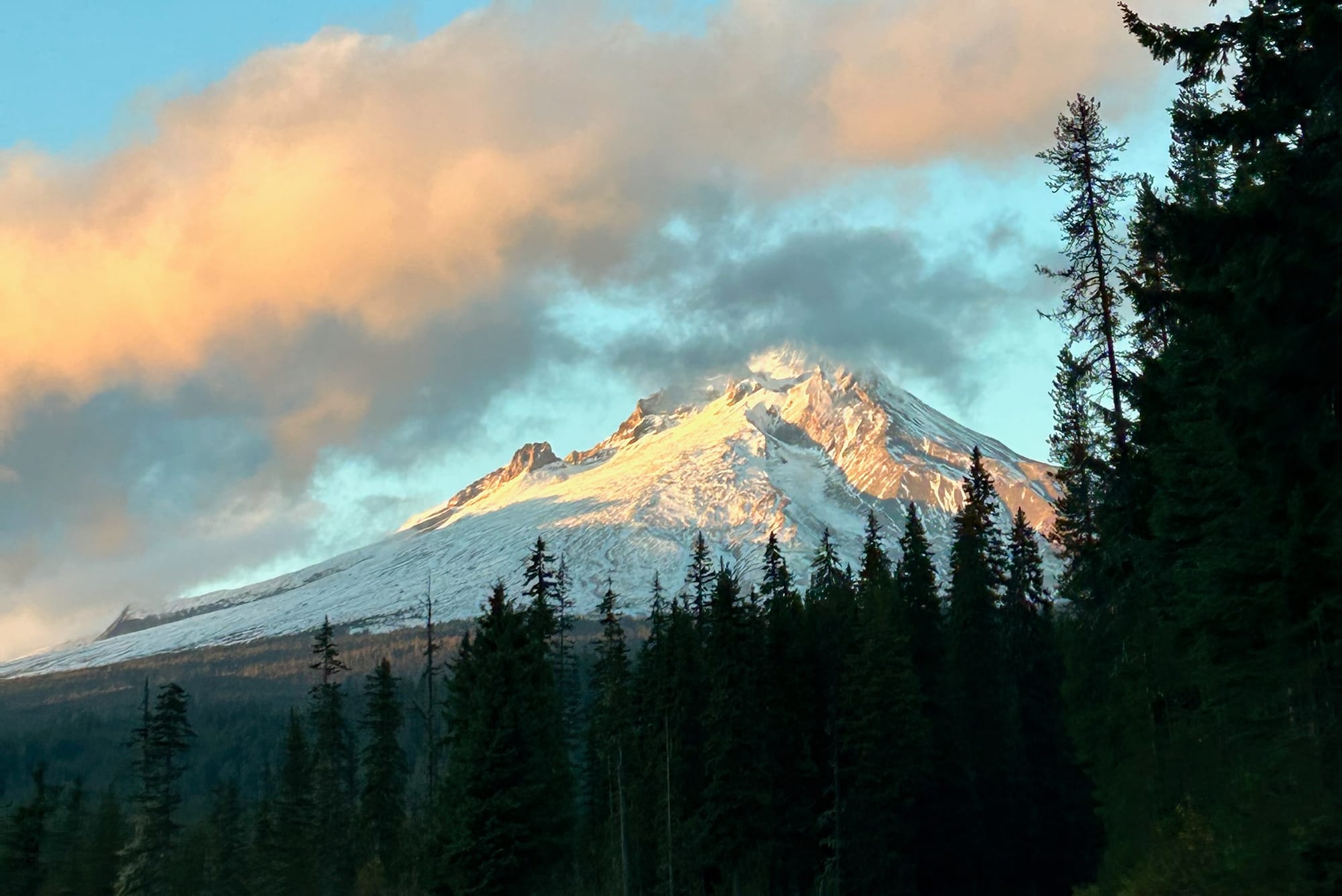 Sunset photo of Mount Hood in Oregon, with sun shining on the side of the mountain, mixed clouds in the sky, and shaded trees in the foreground.