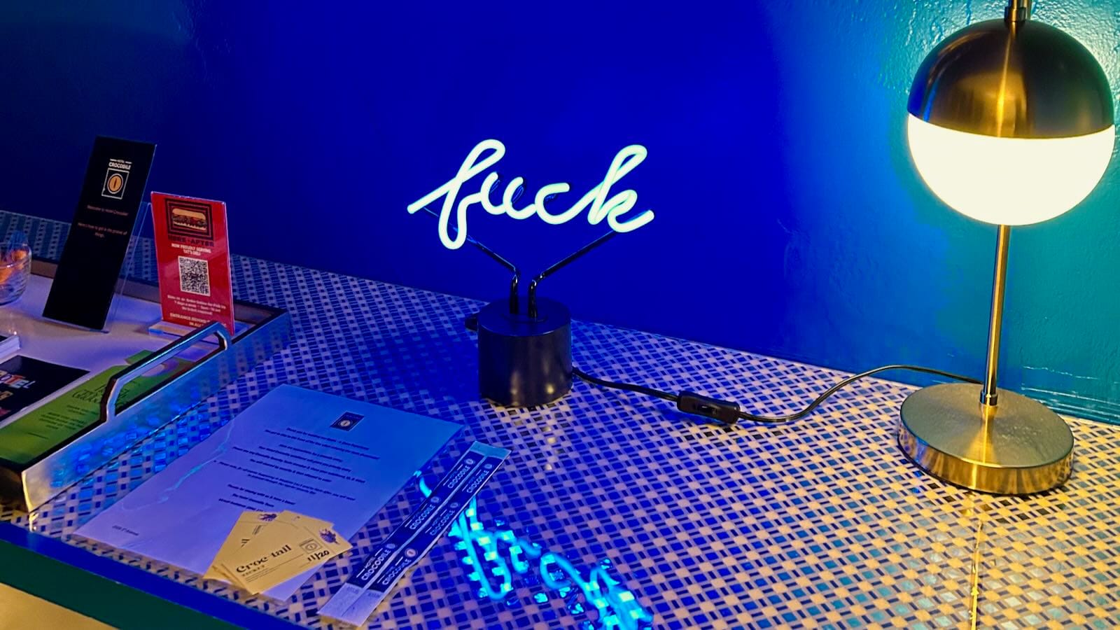 A table in a hotel room at The Crocodile with a neon sign that reads "fuck" in cursive, with a blue wall in the background and "cocktail" drink tickets.